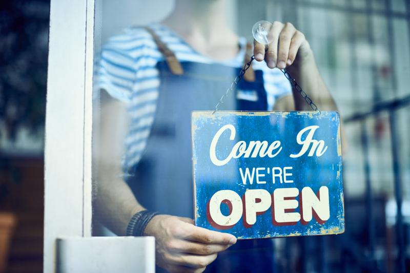 Isang pigura na naka-overalls ang may hawak na asul na karatula sa bintana ng tindahan na may nakasulat na "Come in, we're open"